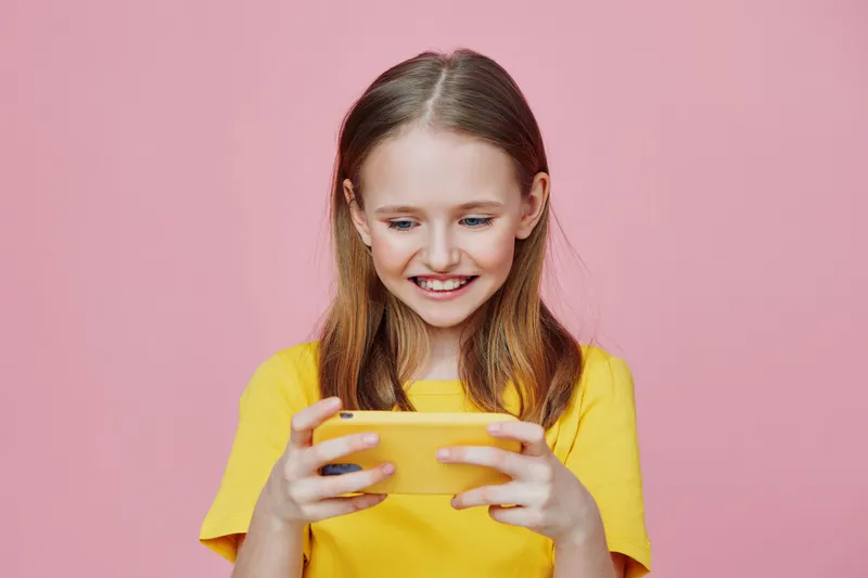 Happy young girl with long hair smiles while playing on her smartphone in a yellow t shirt against a pink background, showcasing joy and engagement in technology