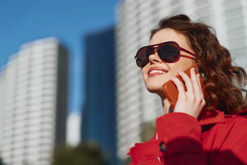 Young woman with curly hair wearing sunglasses and a red jacket smiles while talking on a smartphone outdoors in an urban setting on a sunny day.