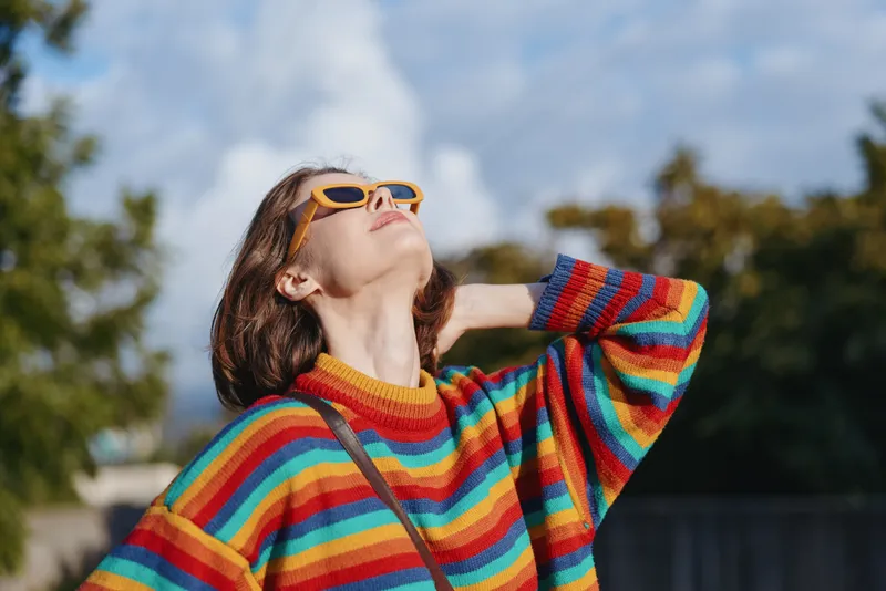 Woman in a colorful striped sweater and yellow sunglasses outdoors, smiling and looking up with short brown hair, casual outfit and crossbody bag, relaxed lifestyle and travel mood.
