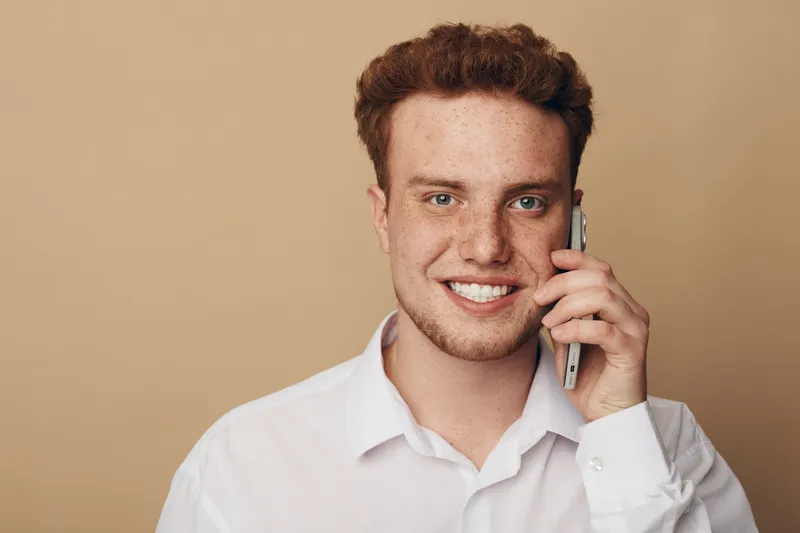 Smiling young man with curly hair wearing white shirt talks on smartphone. Close-up portrait on beige background. Concept of communication, technology, and positive emotions.