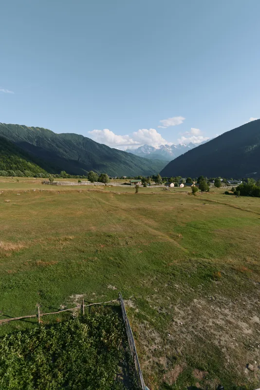 A tranquil rural landscape features an expansive green field bordered by distant mountains under a bright blue sky, with a rustic fence in the foreground.