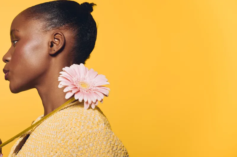 Profile of a young black woman with dark skin wearing a yellow sweater holding a pink flower on her shoulder. Concept of natural beauty, femininity, and floral elegance on yellow background.