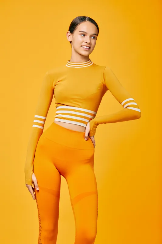Woman fitness model in orange athleisure posing confidently in a vivid studio. Athletic sport vibe with cropped top and high-waist leggings, bright orange backdrop highlighting movement and an active