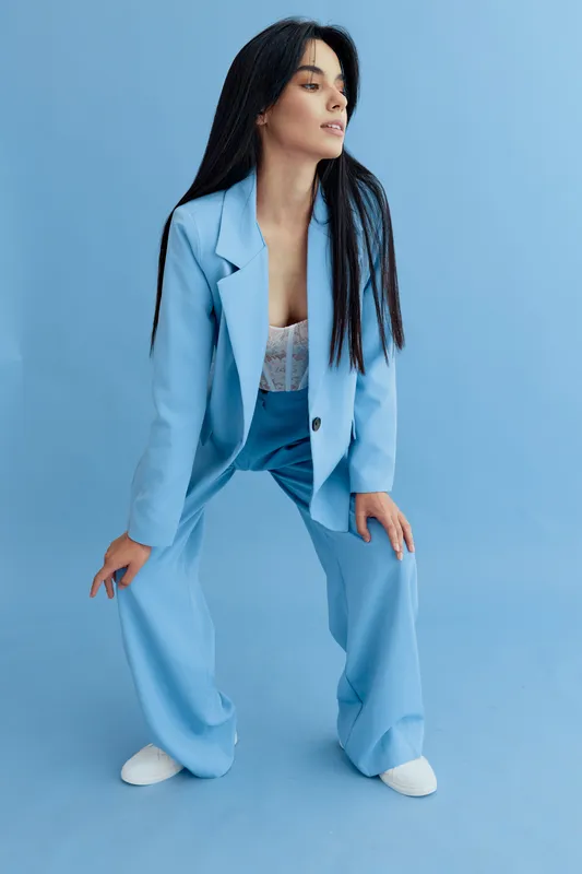 Stylish woman in a light blue pantsuit posing confidently against a matching blue background, showcasing modern fashion with long dark hair and white shoes.