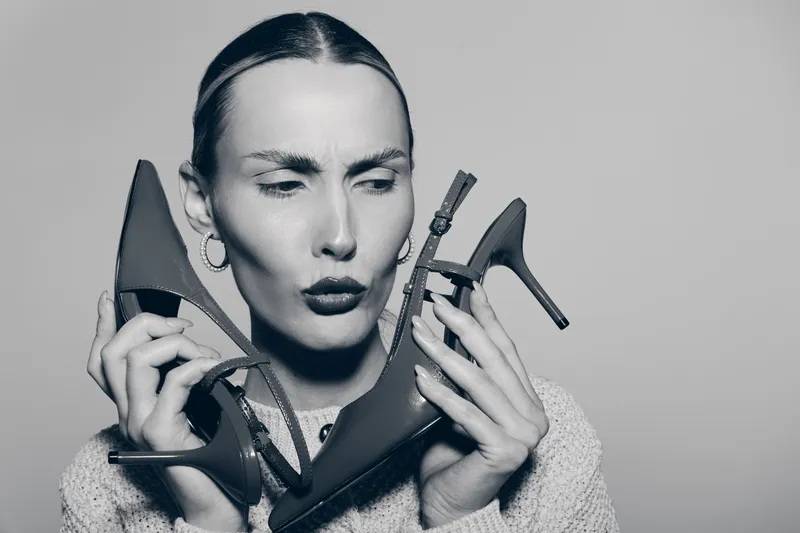 Caucasian woman with pale skin holding two high heels, looking confused and thoughtful. Concept of fashion choice and indecision.