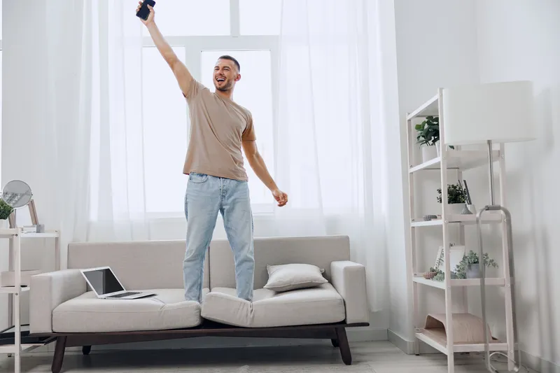 Happy man expressing joy while standing on the couch, celebrating digital detox and embracing emotional minimalism in a bright and airy living room.