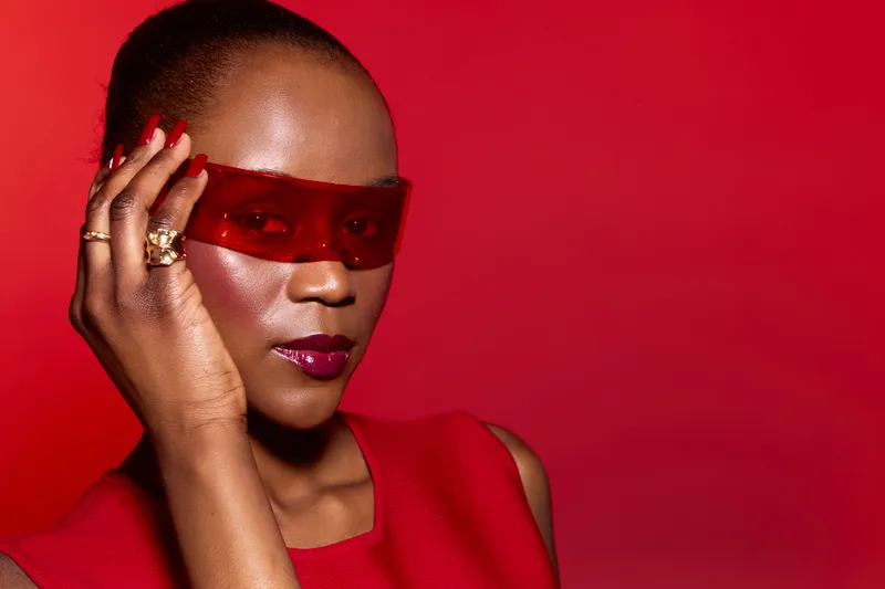 Confident black woman with dark skin wears red sleeveless top and red translucent glasses, posing on vibrant red background. Concept of fashion, style, and modern portrait.