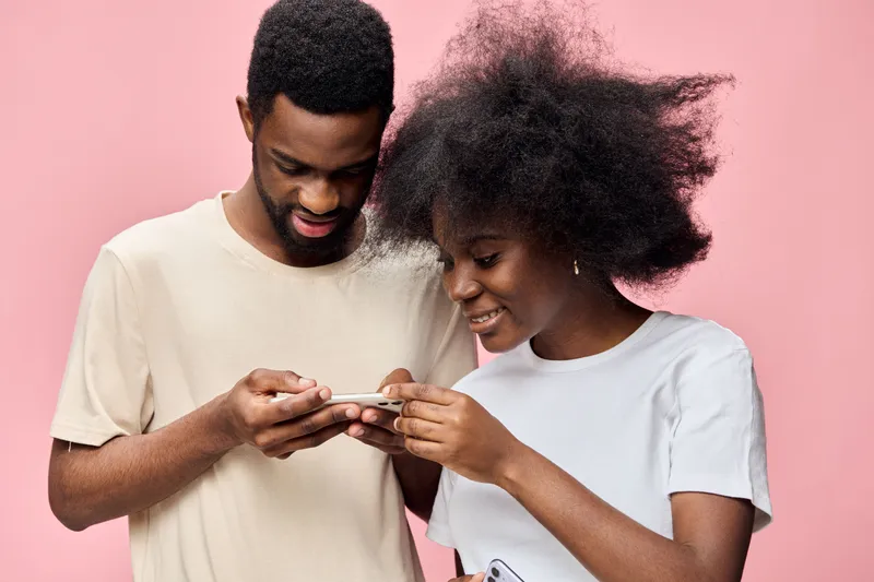 Young African couple enjoying time together, smiling and using a smartphone against a pink background, casual clothing, joyful emotions, modern concept