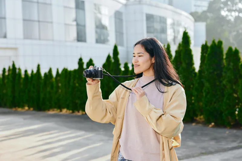 Woman holding a camera outdoors, casual jacket, soft sunlight, focused on framing a shot, greenery and modern architecture in the background, candid photography moment