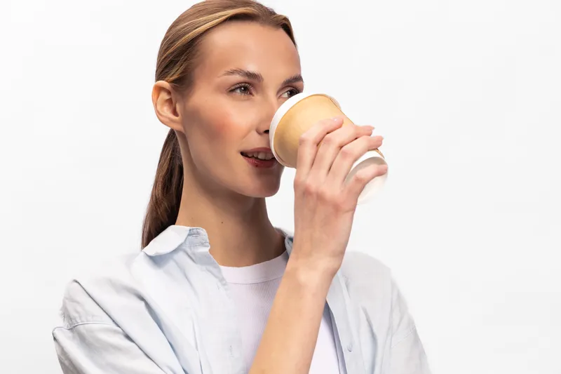 Young woman in casual white shirt drinks coffee from a paper cup, smiling softly. Isolated on white background, concept of morning routine, relaxation, and beverage enjoyment.