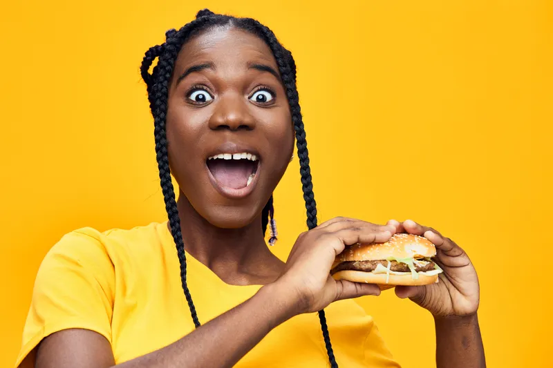 Excited young woman holding a delicious hamburger against a vibrant yellow background, showcasing joy and enthusiasm for healthy eating