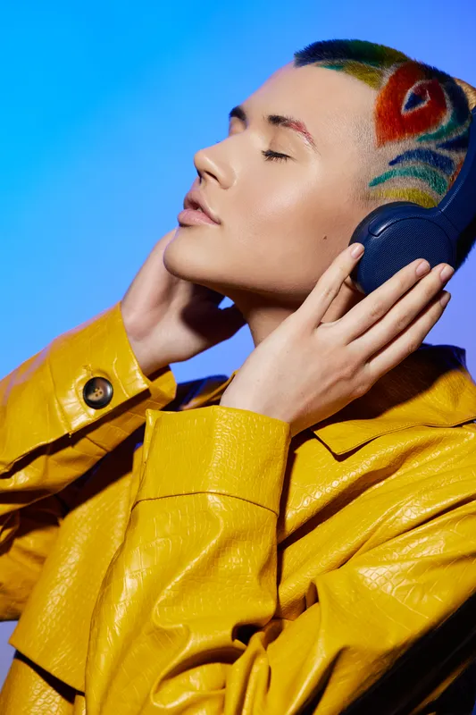 young woman with colorful hairstyle wearing yellow jacket listening to music on headphones, vibrant blue background, artistic expression, modern fashion style
