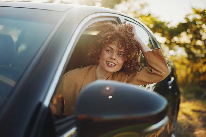 Smiling woman with curly hair leaning out of a car window, warm retro film tones, casual outfit. Lifestyle portrait capturing joy, freedom, and travel vibes.
