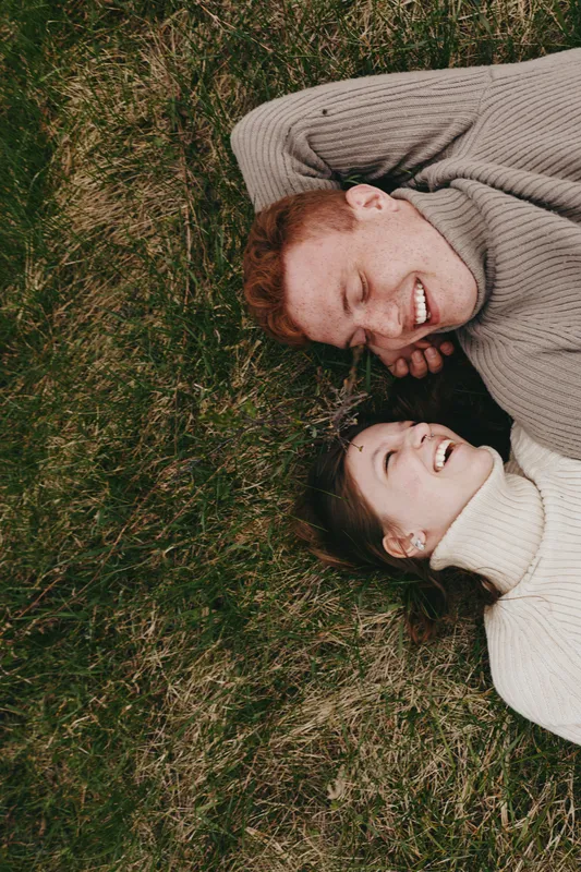 A joyful couple relaxes on a grassy field, smiling and lying close together. A candid lifestyle moment capturing warmth, connection, and comfort in nature, with soft light and casual outfits.
