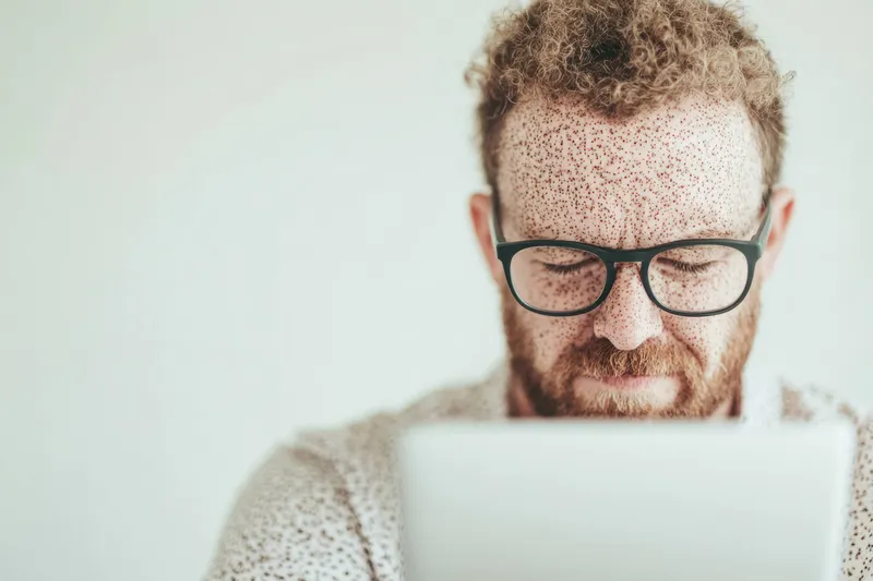 Сосредоточенный мужчина с веснушками работает за ноутбуком, демонстрируя решимость и концентрацию в светлом минималистичном окружении
