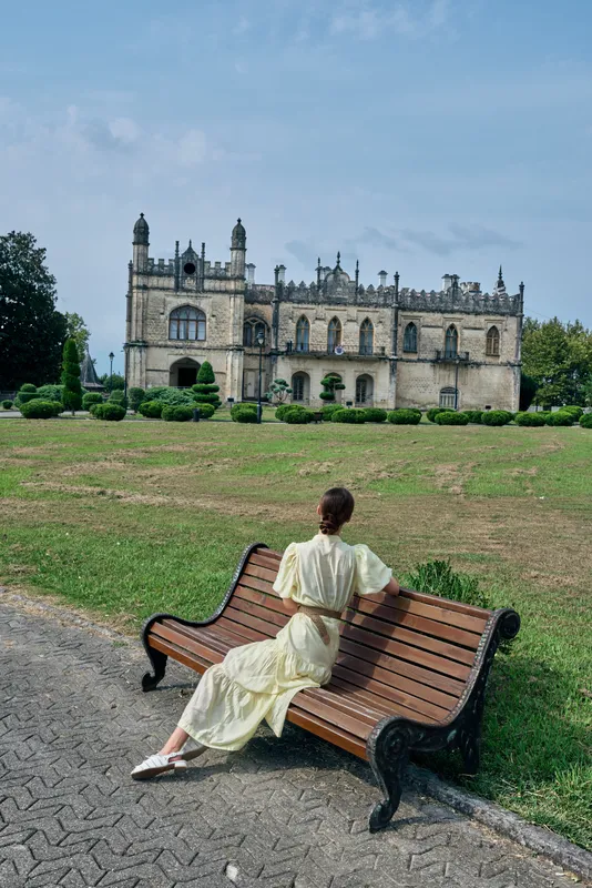 Elegant woman in a light dress sits on a wooden bench in a spacious park, with a grand historic castle in the background, creating a serene outdoor scene.