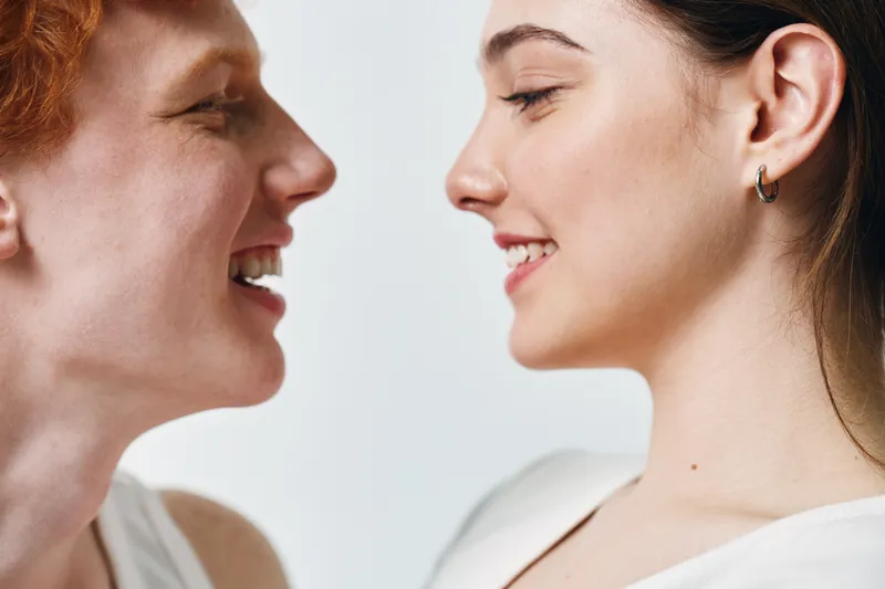 Two young women facing each other, smiling and showing positive emotions with close profile view on white background. Happiness, connection, friendship and diversity concept.