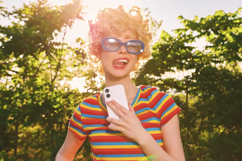 Young woman in a bright striped shirt smiles outdoors while holding a smartphone, sunglasses perched on her nose, sunlit trees framing a lively candid moment, her carefree energy and retro vibe.