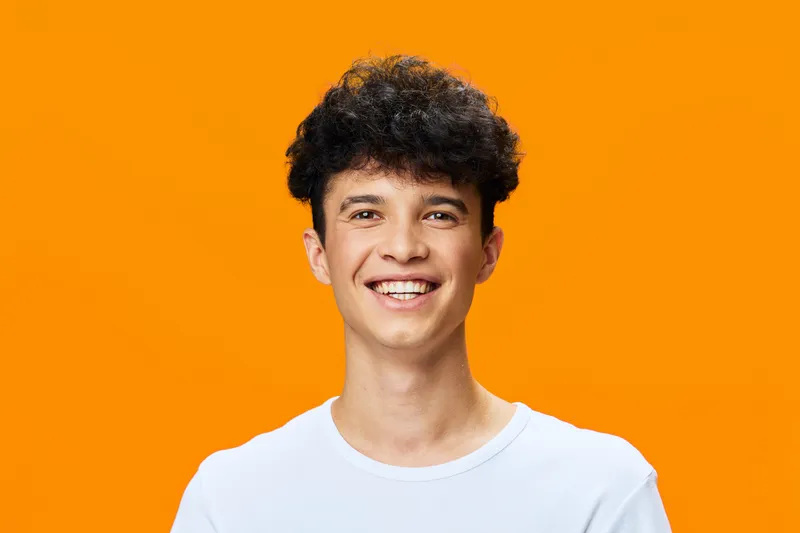 smiling young man with curly dark hair wearing a white t-shirt, joyful expression, bright orange background, casual positive atmosphere, diverse ethnicity