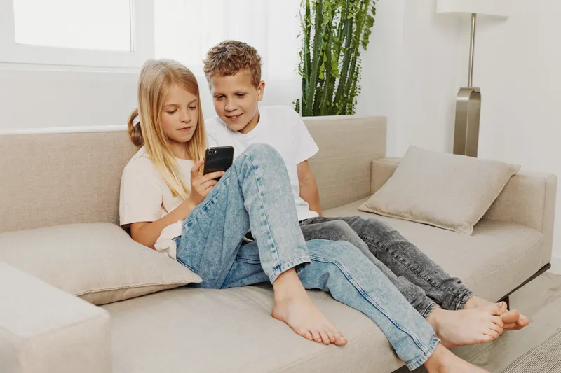 Two children sitting barefoot on a beige couch, using a smartphone together. Bright natural light fills the modern living room with neutral tones and green plants in the background.