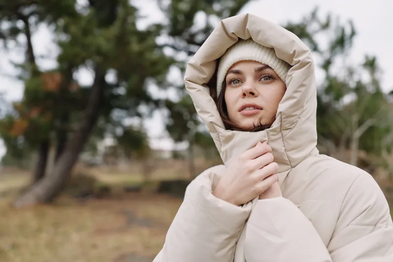 Cold weather portrait of a woman in a beige hooded coat standing outdoors with soft natural light, a calm landscape and a subtle sense of winter fashion and solitude.