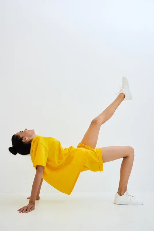 woman exercising in yellow outfit, stretching leg up, wearing white sneakers on white background, fitness pose, active lifestyle