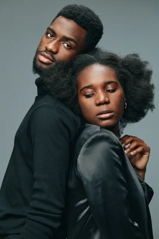 A stylish couple posing back to back, showcasing modern fashion and confidence with natural hairstyles, set against a minimalist gray background