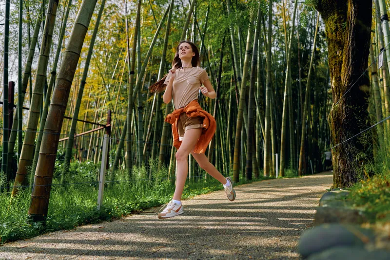 Woman jogging on a forest path through tall bamboo, runner in athletic outfit exercising outdoors, smiling midstride on a sunlit nature trail for fitness and healthy lifestyle.