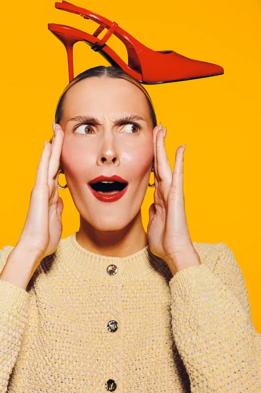Surprised woman with red lipstick holds red high heel shoe on her head against bright yellow background. Concept for fashion, style, creativity, and unexpected ideas.