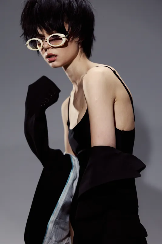 Stylish woman with short black hair wearing sunglasses and black outfit posing against gray background