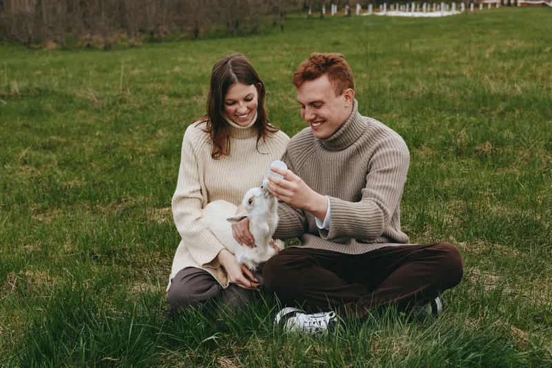 Couple sits outdoors on a field, feeding a goat kid with a bottle, warm interaction and calm mood, relaxed lifestyle scene with natural light highlighting affection