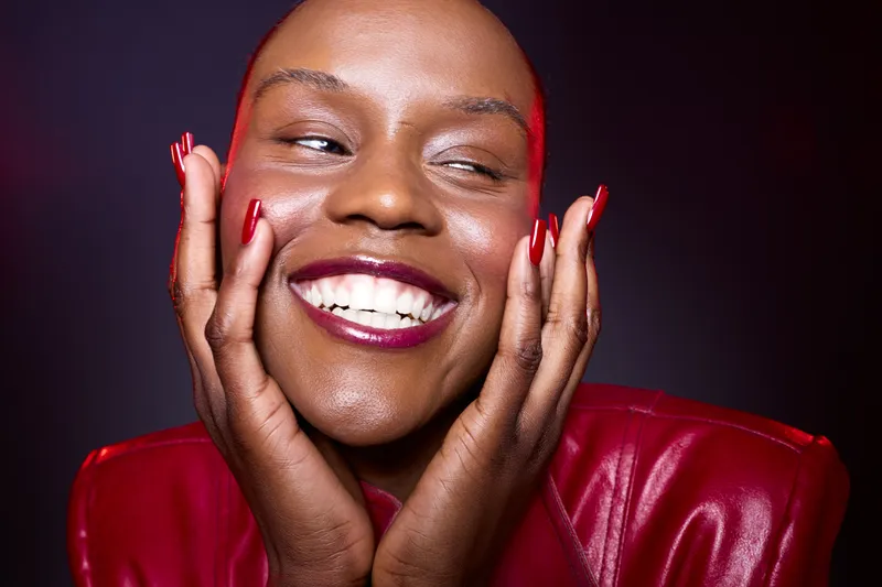 Close-up of happy African woman with dark skin smiling joyfully, hands gently touching face. She wears a red leather jacket and glossy lipstick on a dark red background. Concept of beauty and