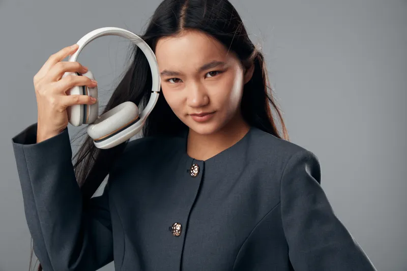Young woman in dark jacket holding wireless headphones for music or technology use against gray background. Portrait of female with unbuttoned suit and modern device for sound entertainment.