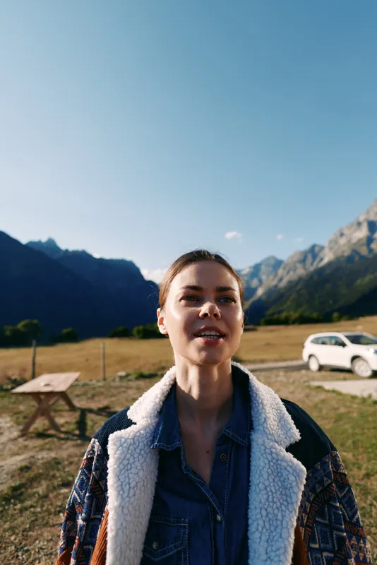 woman mountains outdoor car portrait nature: young female traveler in a shearling denim jacket stands in an alpine meadow by a picnic bench and parked vehicle under clear blue sky.