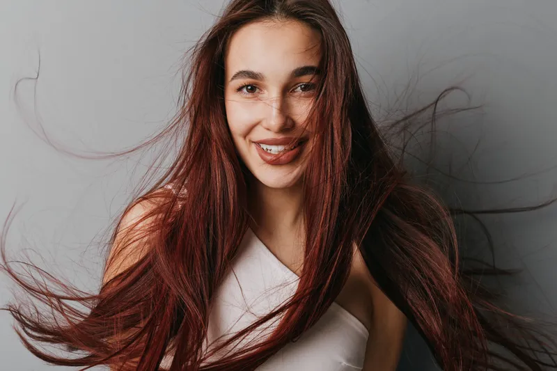 Young woman with long red hair smiling at camera with loose strands flying, wearing white top against gray background, natural look and joyful expression in studio portrait.