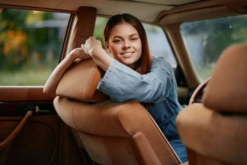 Young female model smiling inside a vintage car, wearing denim dress with natural lighting, showcasing joy, comfort, and casual style in a vibrant outdoor setting.