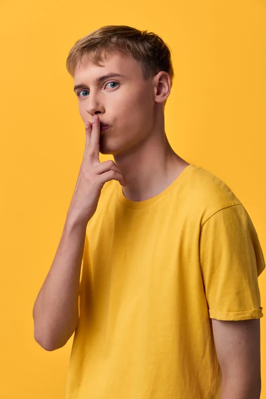 Shushing gesture by a man in a yellow shirt in a studio, conveying silence and secrecy with a calm, minimal mood and soft lighting, clean background, focused expression