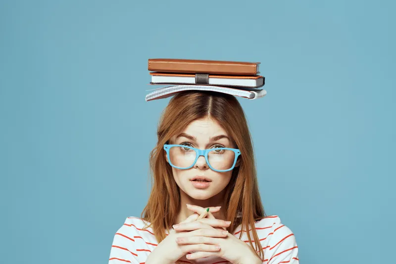 Студентка держит книги на голове на фоне синего цвета, что символизирует научное образование