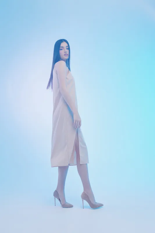 Young woman in beige dress and high heels poses confidently on blue background. Concept of woman beauty fashion and elegance in studio lighting.