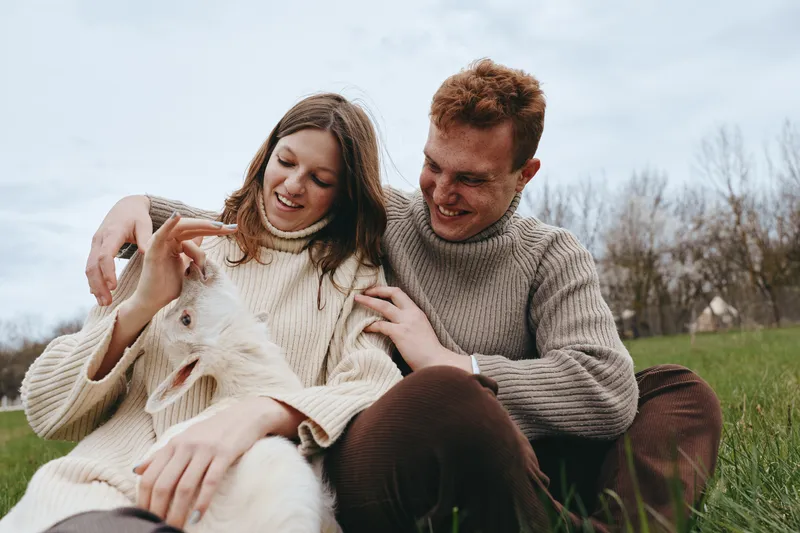A couple smiles as they interact with a friendly goat outdoors in a sunny countryside field, showcasing a lifestyle moment of companionship, warmth, and love for animals in a natural setting.
