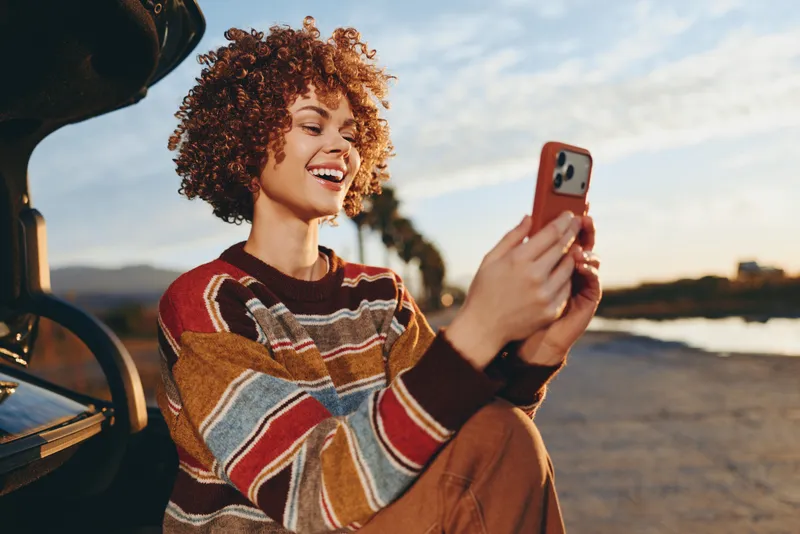 Smiling woman with curly hair, holding a smartphone selfie outdoors, sunlit by water, warm sunset light, casual sweater, relaxed travel lifestyle moment, scenic blurred background