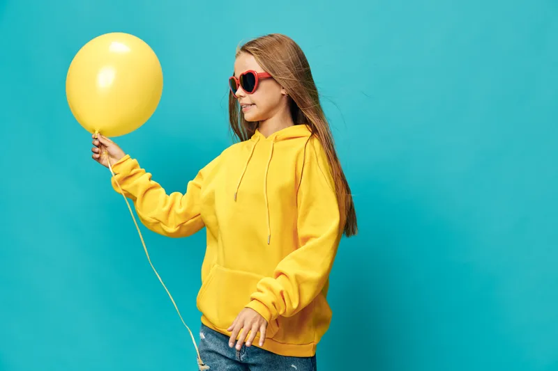 Girl in yellow hoodie and sunglasses holding yellow balloon, posing on blue background with casual style and relaxed expression for birthday or party concept.