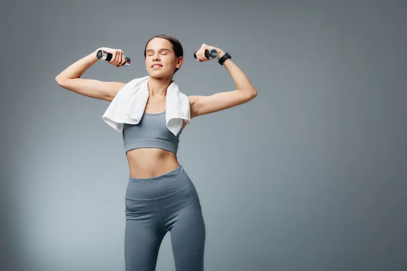 Fitness woman in athletic wear flexes arms with dumbbells, towel around shoulders, showing strength and confidence during a focused workout in a clean studio.