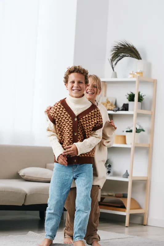 Two smiling children embrace indoors wearing knitted sweaters, bathed in warm natural light. A cozy living room with sofa and soft decor frames a cheerful moment.