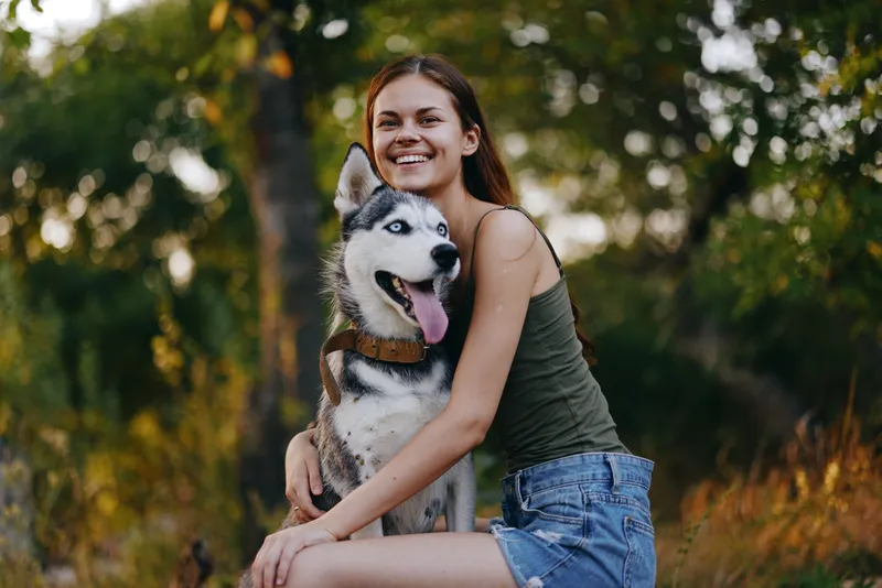 Радостная женщина с собакой породы хаски улыбается, сидя на природе во время прогулки с собакой на поводке, на заднем плане осенний пейзаж