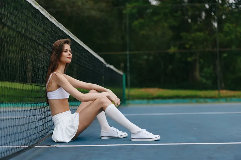 Young athletic woman in sportswear sitting on a tennis court, exuding confidence and determination, surrounded by lush greenery