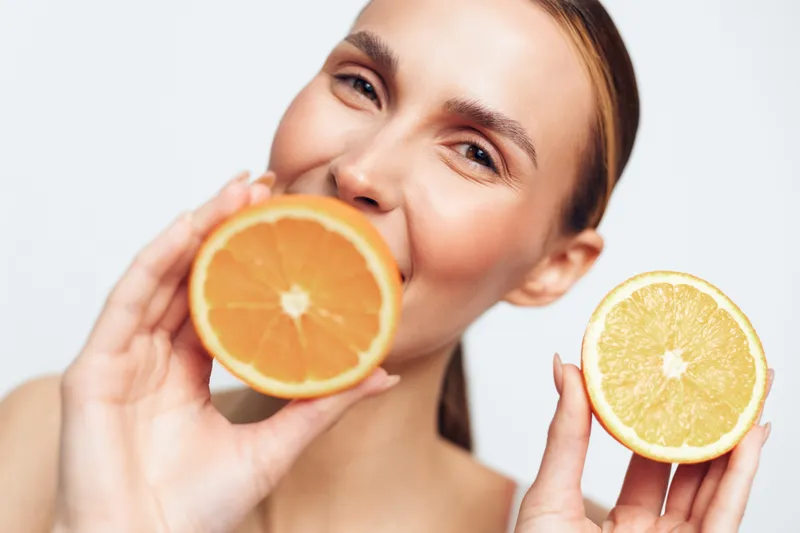 Young woman holds two halves of fresh orange near face smiles showing healthy skin and natural beauty isolated on white background closeup portrait with citrus fruit.