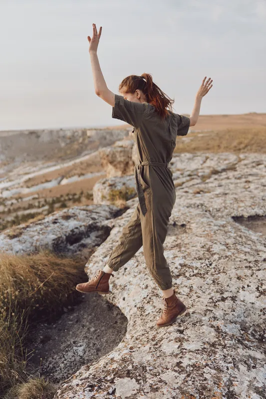 Женщина в зелёном комбинезоне стоит на вершине скалы с распростёртыми руками на фоне величественного пейзажа