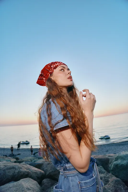 Lifestyle woman in modern boho-western denim street style with long hair and red bandana, captured in warm film color tones at the beach during sunset time.