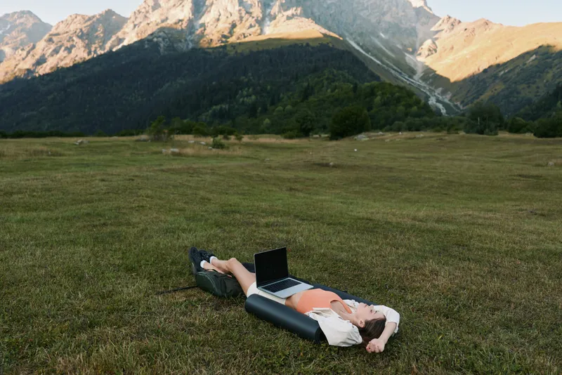 A person lies on a grassy field with a laptop beside them, enjoying sunlight in a mountainous landscape. Calm and focused, they work remotely outdoors, embracing nature during a peaceful break from